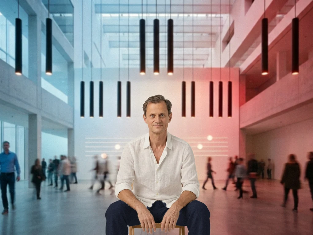 Klavierpädagoge Michael Köllner in weißem Leinenhemd und dunkler Hose sitzt ruhig auf einem Hocker in einer hellen Halle, über ihm schwarze Leuchtröhren in Gruppen wie die schwarzen Tasten eines Klaviers, hinter ihm Lichtlinien und Menschen in Bewegung